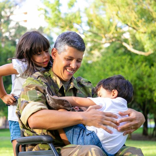 Happy disabled military dad walking with two children in park. Girl holding wheelchair handles, boy resting on dads lap. Veteran of war or disability concept